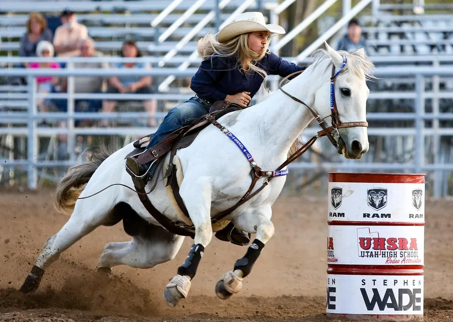 A young rider in a white hat on a grey horse leaning hard into a turn around a red and white barrel, dust kicking up behind