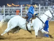 A rider on a grey horse turning tight around a blue barrel under arena floodlights, sand spraying from the horse's feet