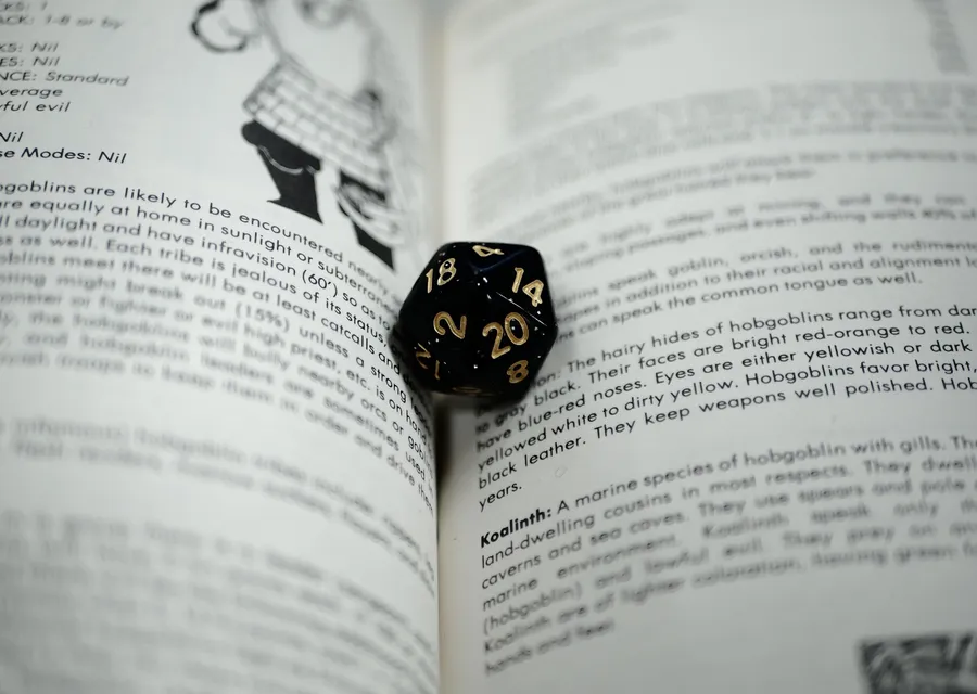 A black twenty-sided die resting in the gutter of an open rulebook, the page describing hobgoblins